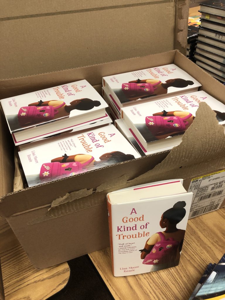a box of hardcover books with the title that reads A Good Kind of Trouble and shows an illustration of a 12 year old Black girl wearing a pink backpack with patches on it